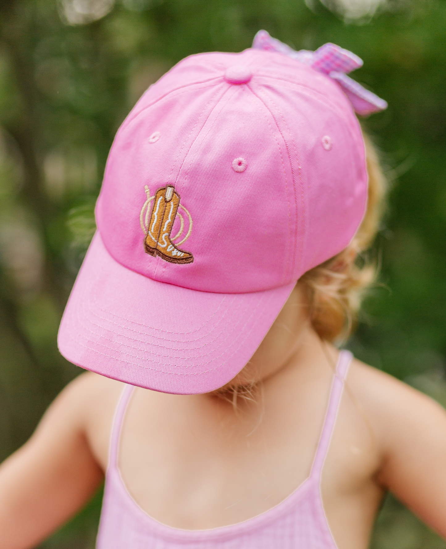 Girl wearing magenta baseball hat with cowboy boot embroidery and bow on the back