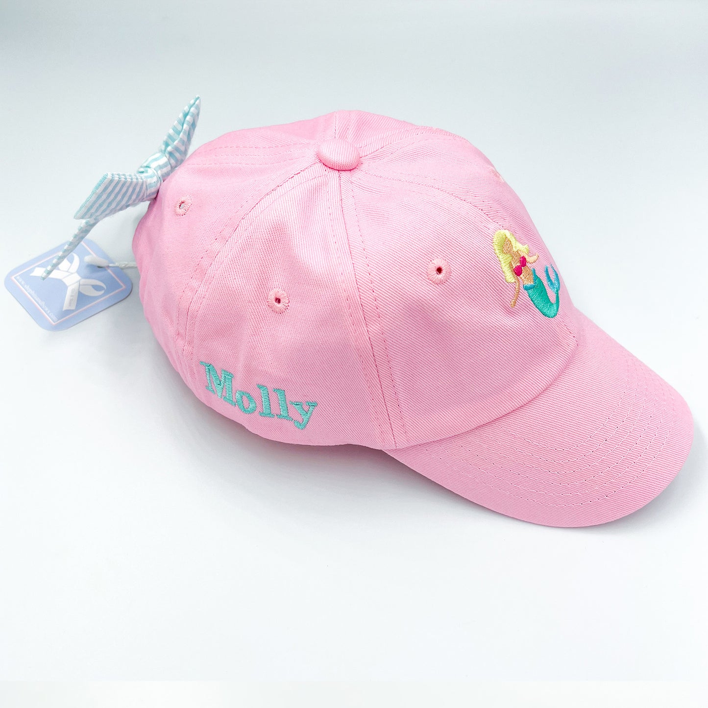 Pink baseball hat with mermaid embroidery on the front, a bow on the back and a name embroidered on the side