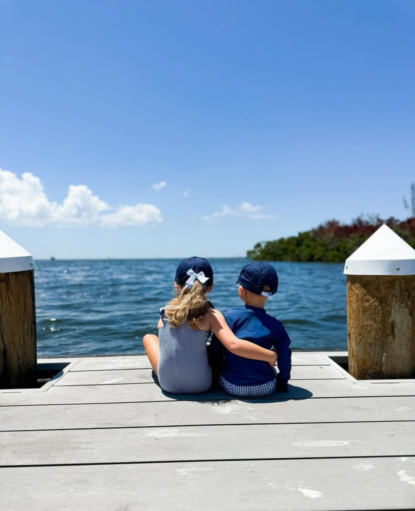 Little boy and girl sitting on a dock wearing coordinating navy blue baseball hats