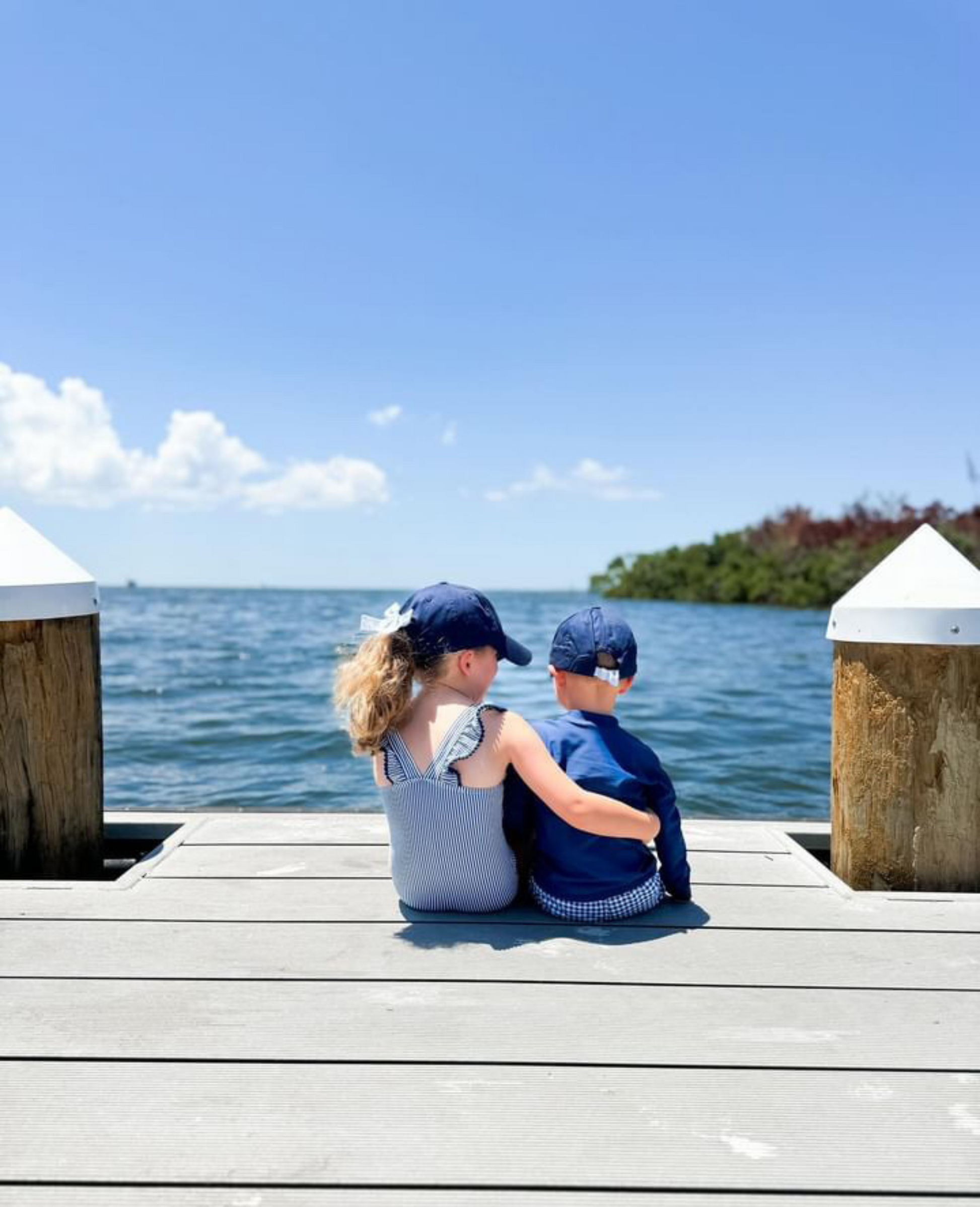 Little boy and girl sitting on a dock wearing coordinating navy blue baseball hats