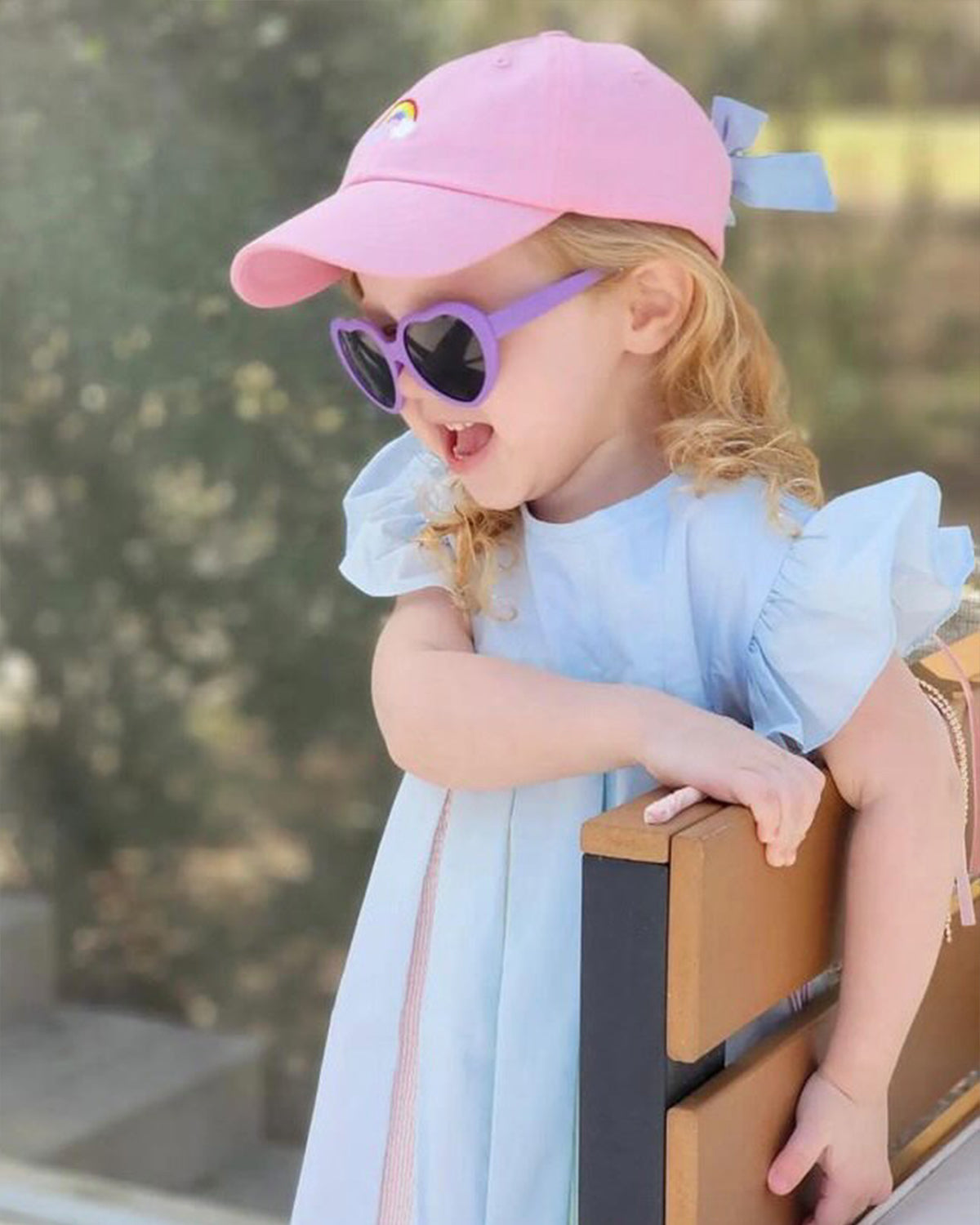 Little girl wearing a pink baseball hat with rainbow embroidery and a bow on the back