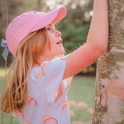 Girl wearing a pink baseball hat with rainbow embroidery and a bow on the back