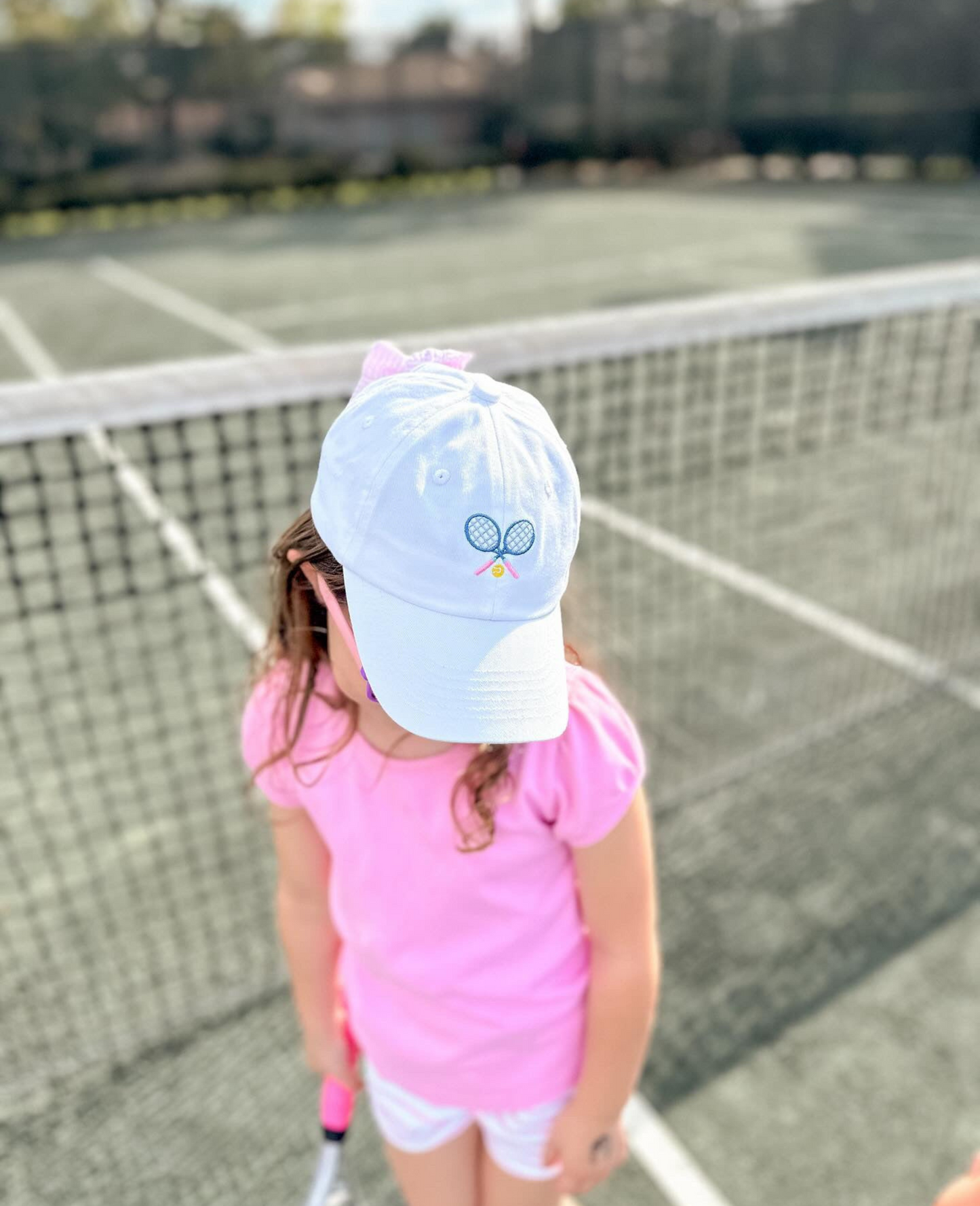 Girl wearing a white baseball hat with tennis racket embroidery and a bow on the back