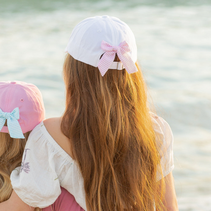 Girl wearing a white baseball hat with a pink and white seersucker  bow on the back